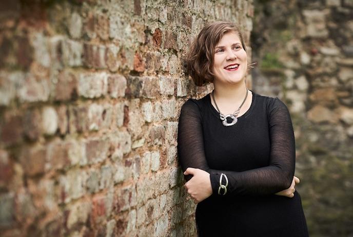 A person wearing a black dress and silver jewellery leans against a multi-coloured brick wall.