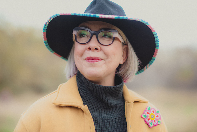 Portrait of Rebecca Hass smiling, wearing a colourful hat and glasses