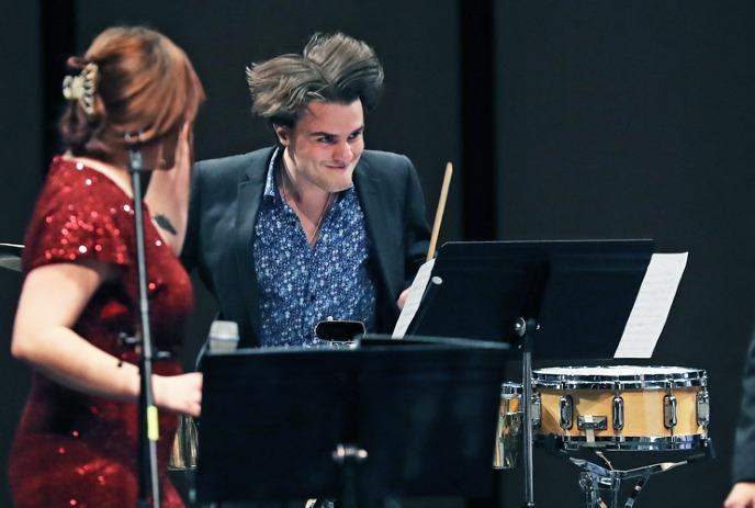Singer in red dress holding a mic with a drummer playing with an expressive stage moment under a dark background.