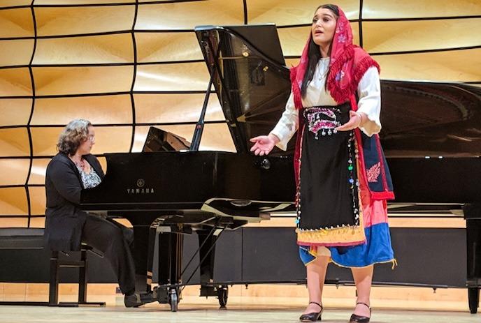 A person wearing cultural clothing sings on stage while a pianist plays a grand piano in the background.
