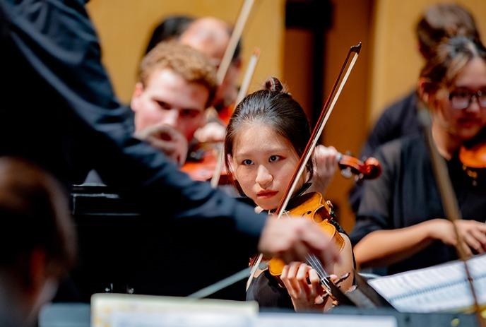Young violinist watches conductor; orchestra in formal black plays under warm lights in concert setting.