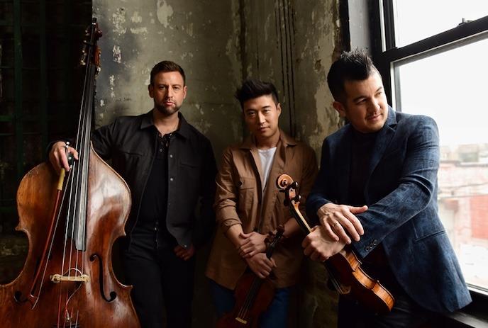 Three artists stand together with their instruments while looking at the camera.