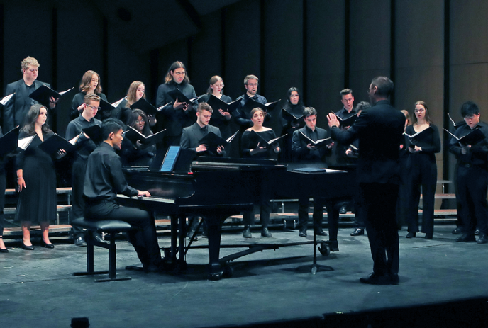 A choir dressed in black performs on stage, accompanied by a pianist.