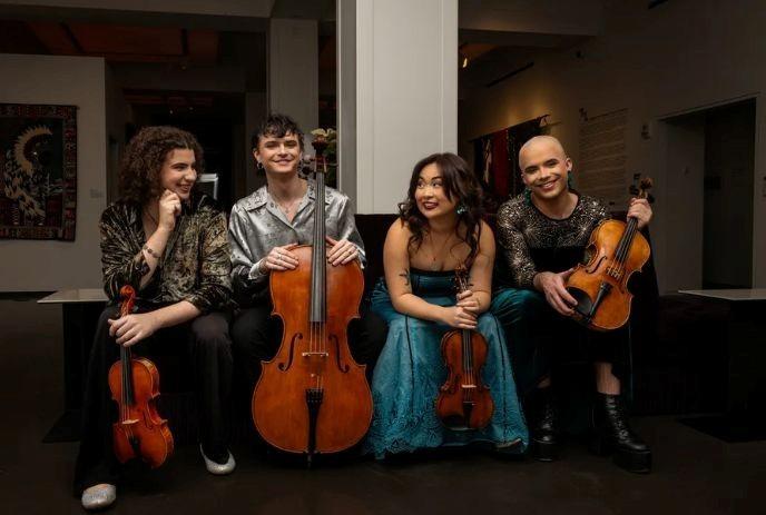 Four diverse musicians in sparkling attire sit with violins, viola, and cello in an art-filled room.