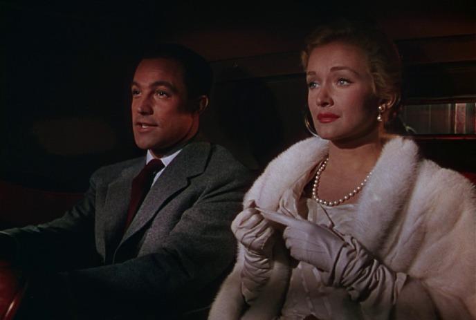 A man in a suit and a woman in a white fur coat with gloves and pearls sit together in a car.