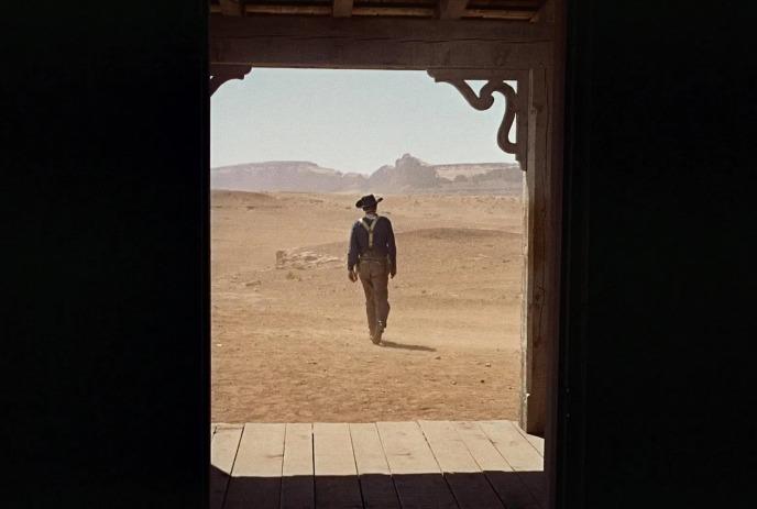 Lone cowboy walking in the desert being watched by someone from a doorframe.