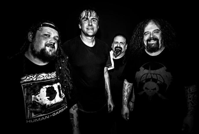 Four men of the band Napalm Death wearing black t-shirts stand closely together, smiling in a dimly lit setting.