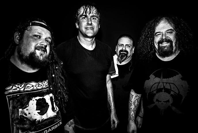 Four men of the band Napalm Death wearing black t-shirts stand closely together, smiling in a dimly lit setting.