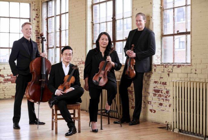 String quartet in bright loft holding violins viola and cello against rustic brick and window backdrop