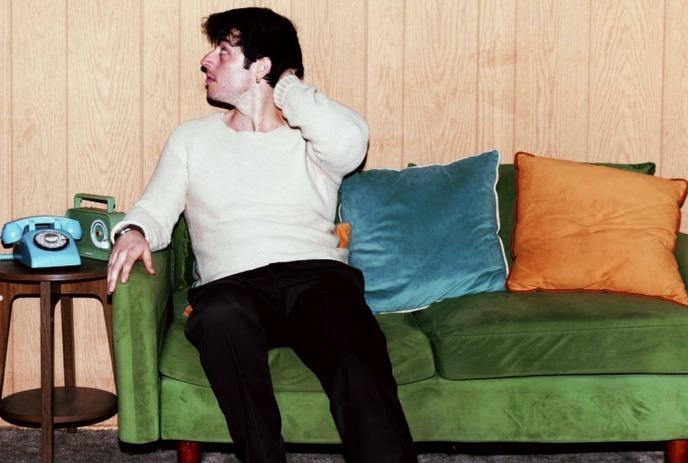 A man in a white sweater sits on a green couch with colorful pillows, looking to his right..
