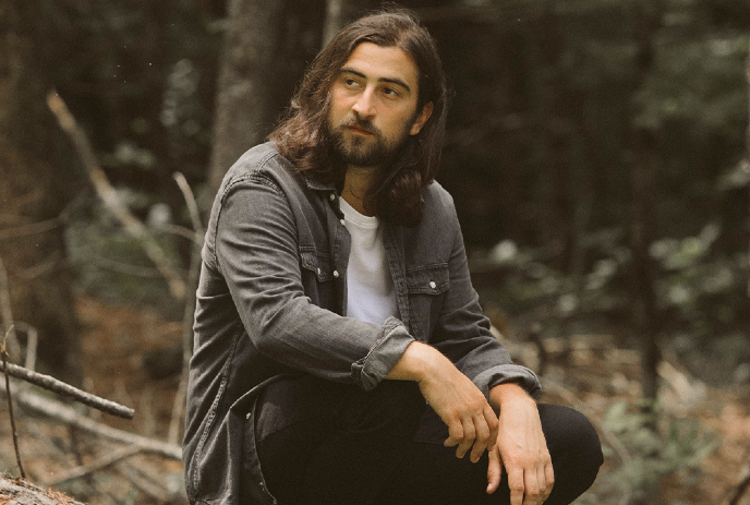 Noah Kahan sits in a forest, wearing a gray jacket over a white shirt.