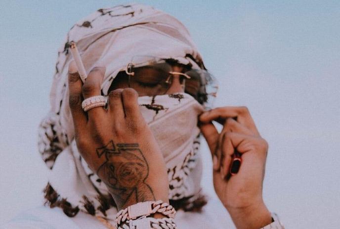 NORTHSIDEBENJI in white, scarf on, holding a cigarette near his tattooed hand with jewelry, against a clear sky.