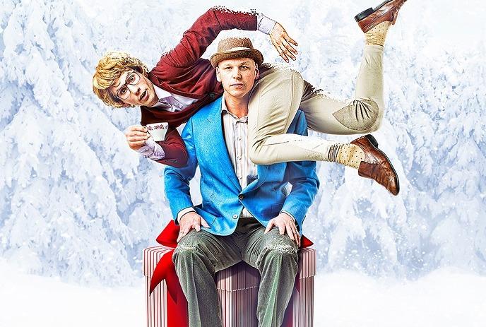 Two people pose playfully in snowy woods, one draped over the other's shoulders while seated on a wrapped gift box.
