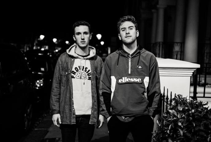 Two young men of Cleopatrick stand on a dimly lit street at night. Both wear casual hoodies, with one in a denim jacket.