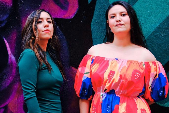 Two women pose in front of a vivid mural; one wears green, the other a floral dress with bold colors.