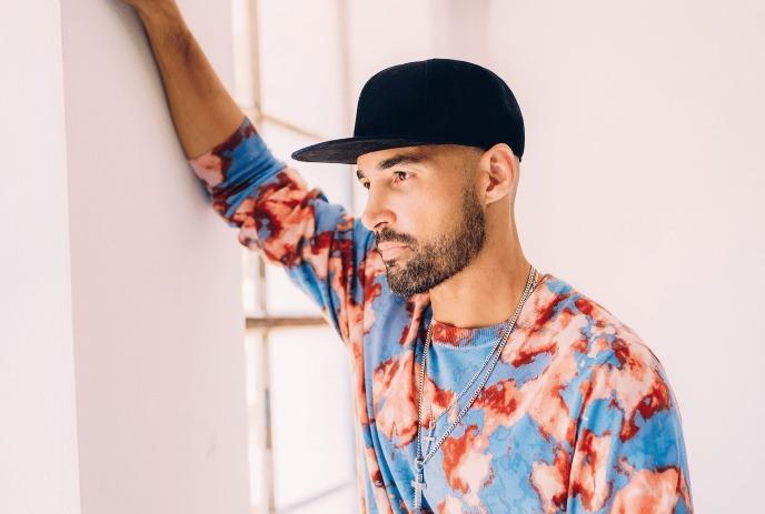 OFFAIAH leans against a wall wearing a black cap, a tie-dye sweater, and layered necklaces.