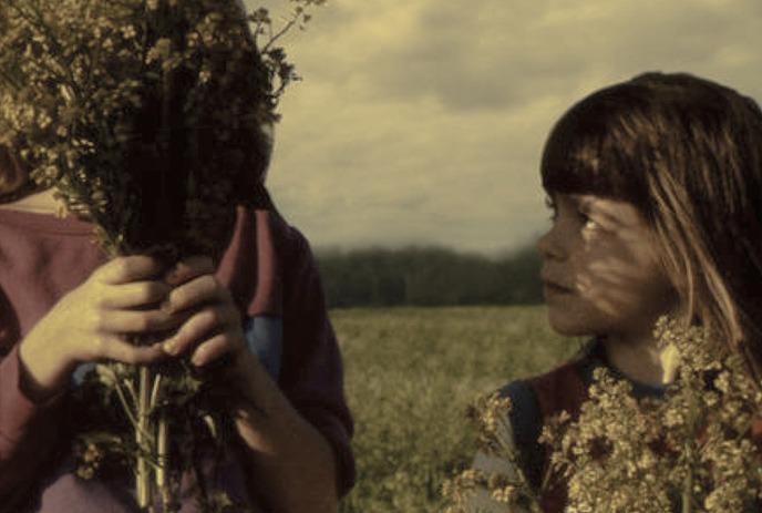 Two kids in a grassy field hold wildflowers; one hides their face while the other smiles softly.