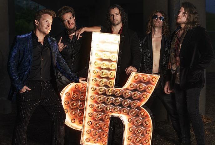 Five artists pose near lit metal OK sigh while singer in cap performs with mic.