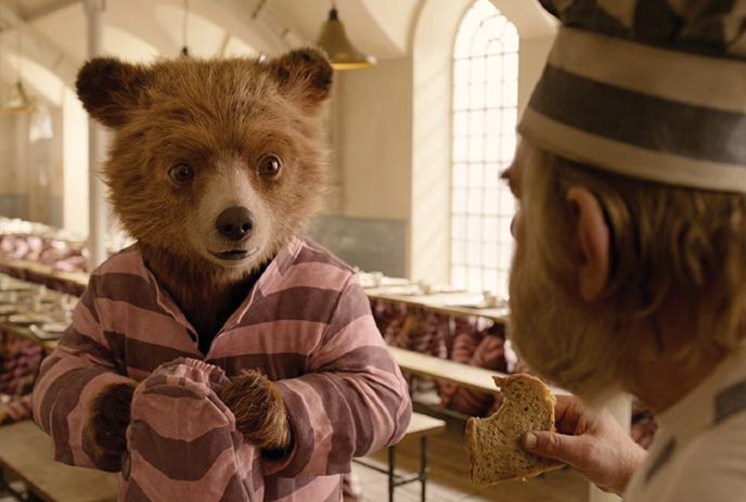 Paddington Bear looking at another person eating a sandwich.