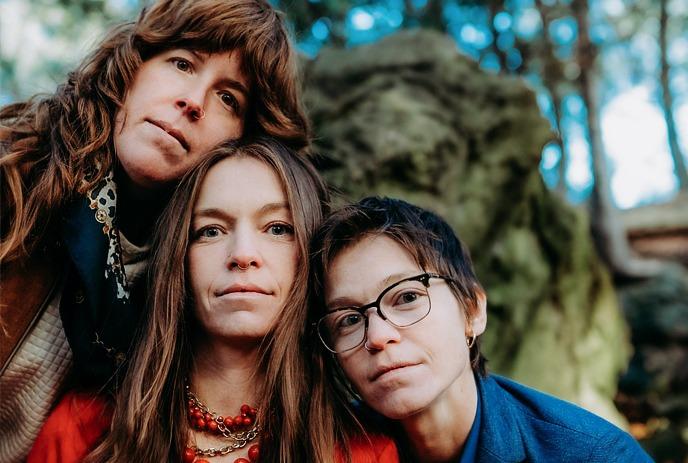 Three artists posing closely together, outdoors with trees and rocks in the background..