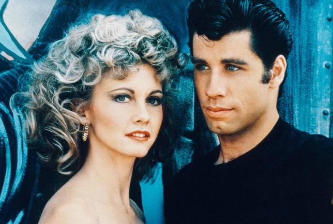 Two people stand close against blue backdrop, one with blond curls and lipstick, one in dark shirt.