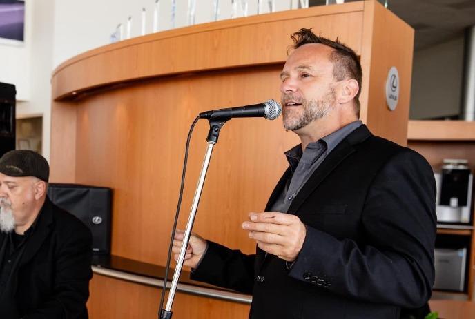Two men in suits, one singing into mic with wooden backdrop.