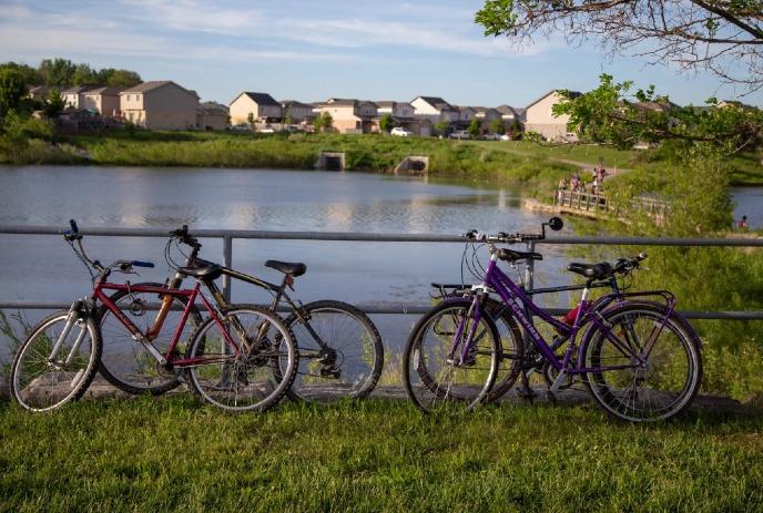 Red and purple bikes rest against railing by calm pond with houses trees and bridge in background