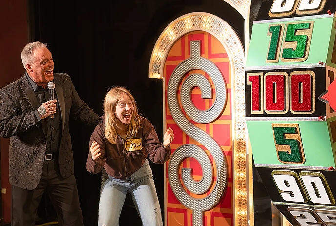 Giant Plinko game board on stage with three people celebrating, arms raised, in a colorful set.