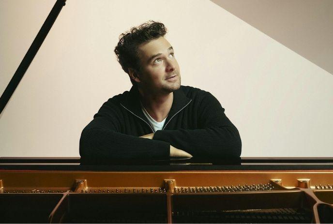 Person seated at grand piano rests arms on edge while gazing upward against softly lit calm background setting.