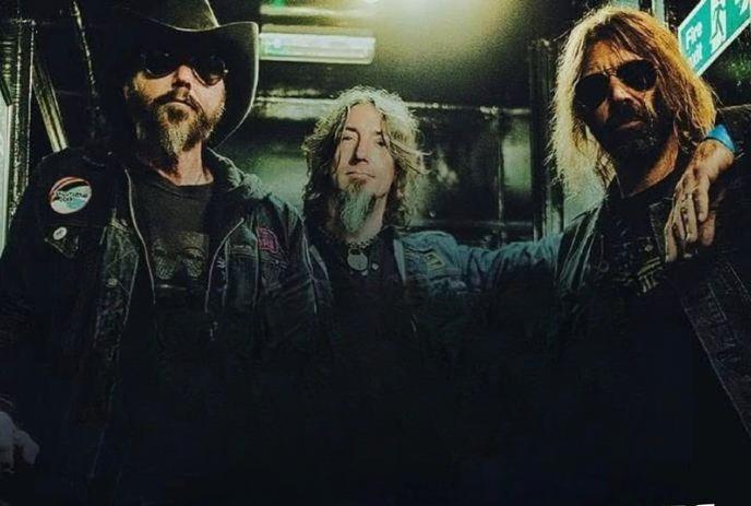 Three artists in dark jackets and sunglasses standing together in dim industrial setting with green exit sign.