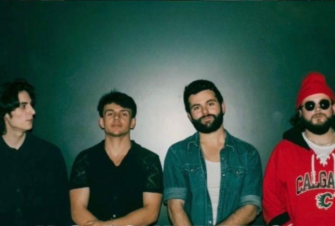 Four artists standing together against a dark green wall wearing casual clothing with varied styles and poses.