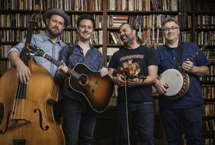 Four musicians stand before bookshelf holding bass guitar fiddle and banjo in warm indoor library setting.