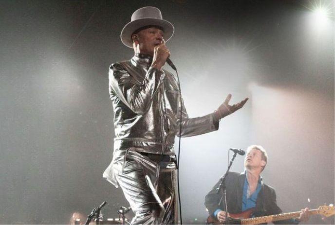 Singer in silver suit and top hat performs on lit stage with guitarist behind amid bright beams.