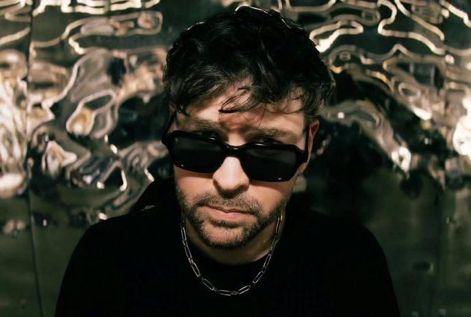 Kyle Watson in dark sunglasses and chain stands before metallic wavy backdrop creating modern abstract.