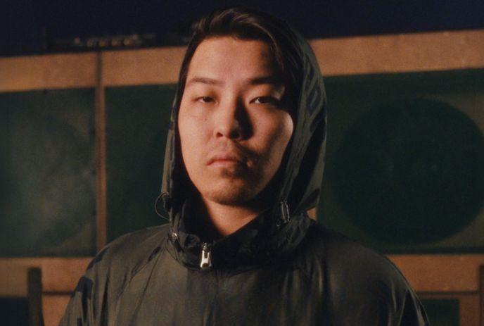 Person in dark hooded jacket stands before warm lit green paneled structure creating cinematic mood.