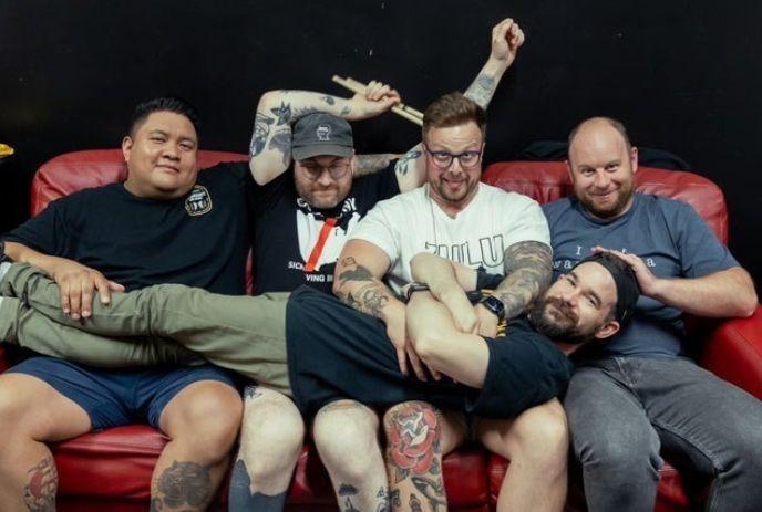 Five friends on red couch with one lying across laps as others pose playfully against dark backdrop.