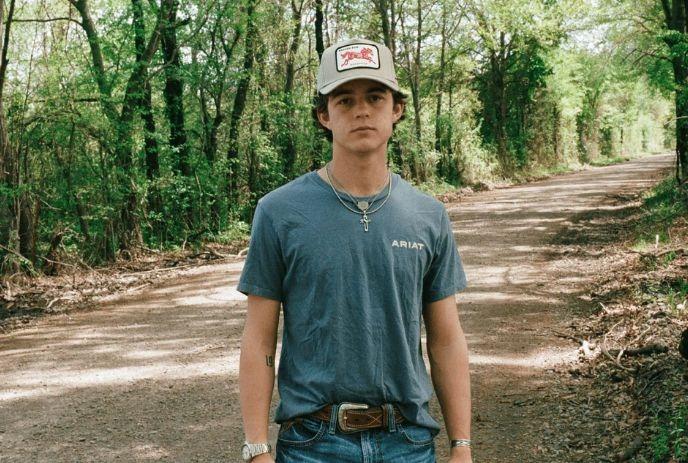 Waylon Wyatt in cap stands on dirt road amid tall green trees creating calm outdoor scene
