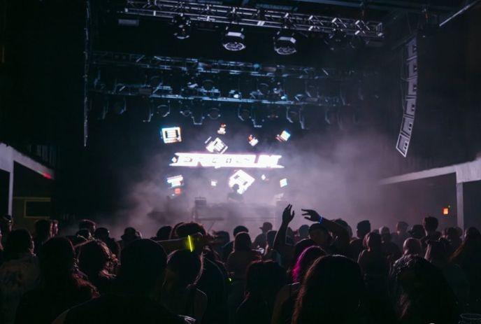 Crowd faces bright stage with lights and fog as people raise hands in energetic live music scene.