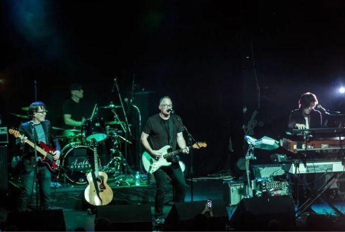 Four musicians play bass drums guitar and keys on dim stage with green blue lights and acoustic guitar nearby.