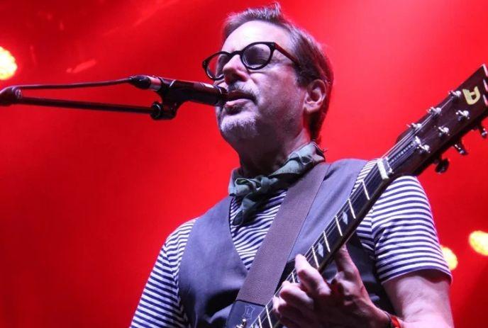 Musician in glasses sings and plays electric guitar under vivid red stage lights with lively concert energy.