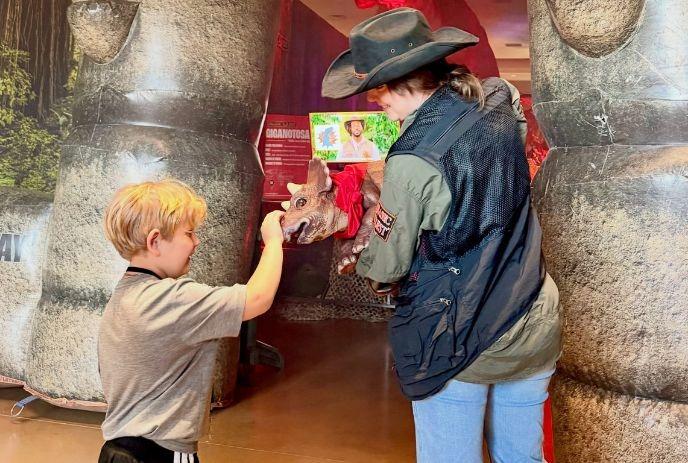 Child touches small dinosaur puppet held by costumed handler near large themed inflatables indoors.