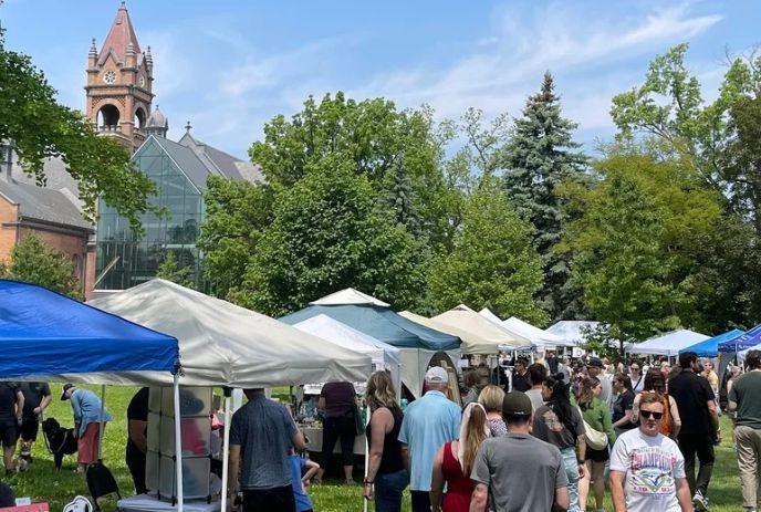 Crowds browse rows of tents on sunny lawn as tall brick clock tower rises behind market scene.