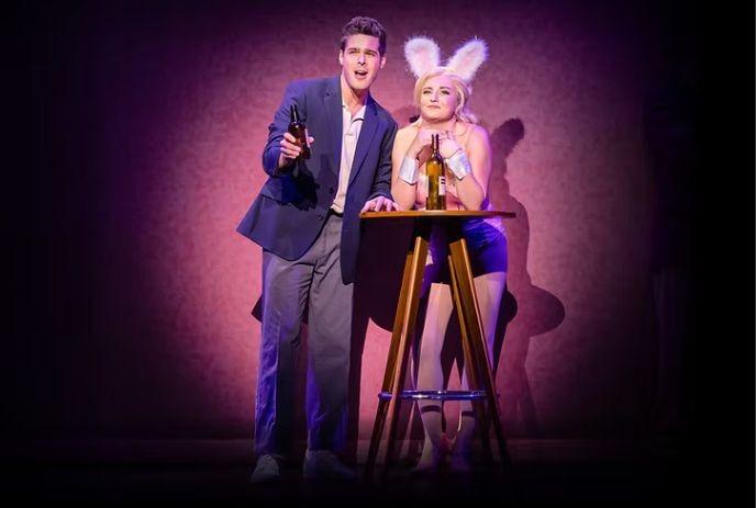 Two performers onstage one in suit holding bottle and singing other in bunny costume leaning on table under purple light.