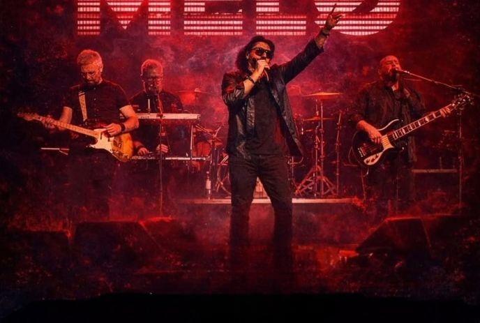 Band performs on red lit stage as singer raises arm with guitarists flanking and drums behind.