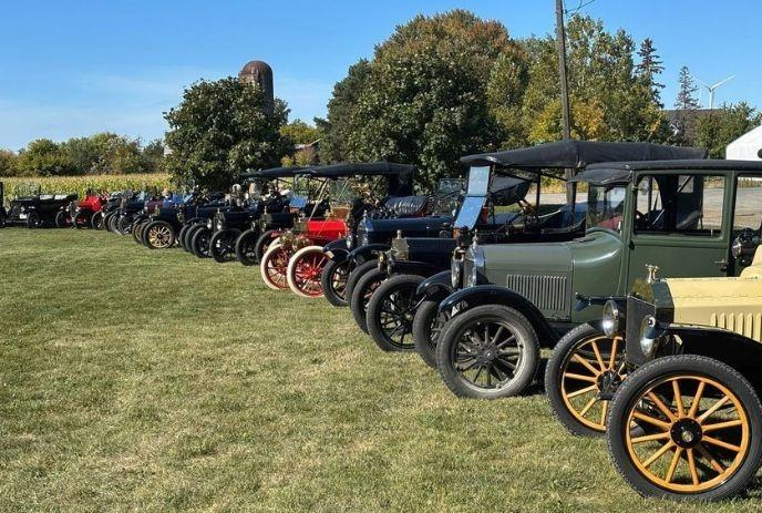 Row of vintage cars parked on grassy field with autumn trees and silo behind under clear sky.