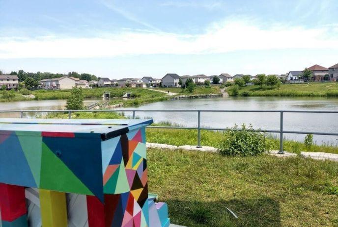 Colorful geometric box stands by pond with houses trees and railing in bright suburban setting.