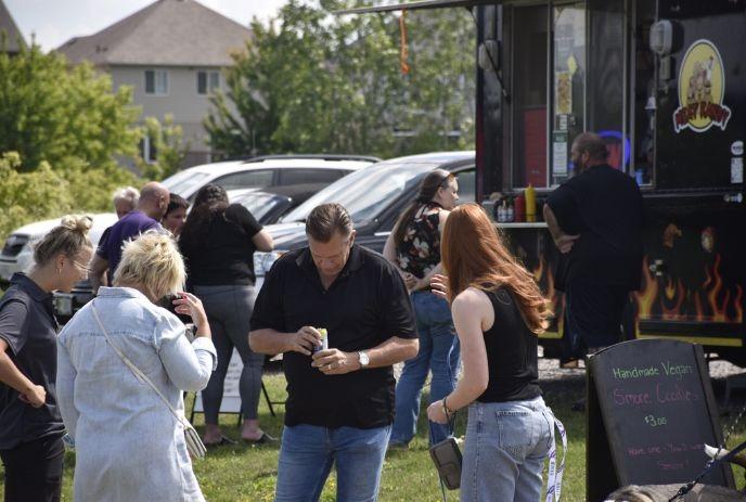 People gather by food truck on grassy area with sign as cars and houses sit in suburban background.