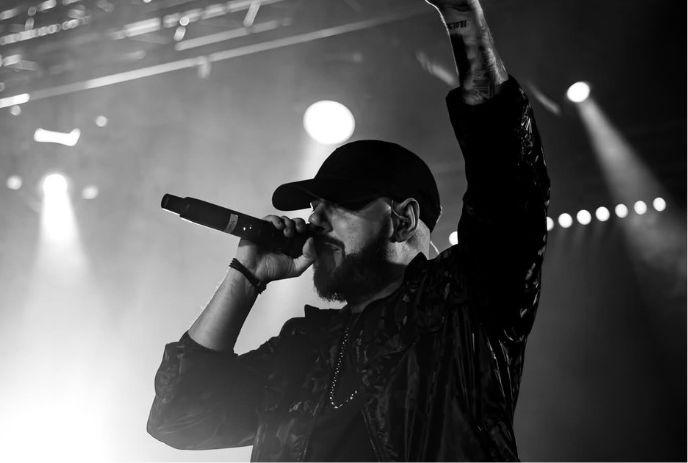Performer raises arm while singing on stage with bright spotlights in dramatic black and white scene.