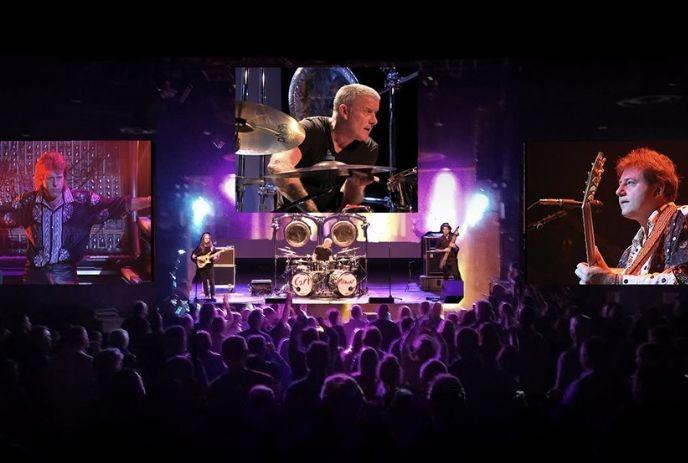 Vibrant rock concert scene with Carl Palmer on drums, purple?blue lights, and crowd energy captured.