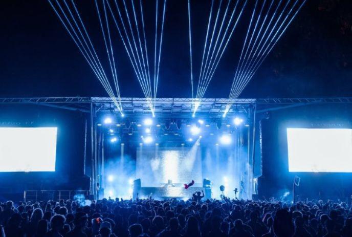 Blue?lit night concert with DJ on stage, crowd cheering under beams of light and glowing screens.